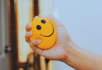 a hand squashing a yellow stress ball with a smiley face on it